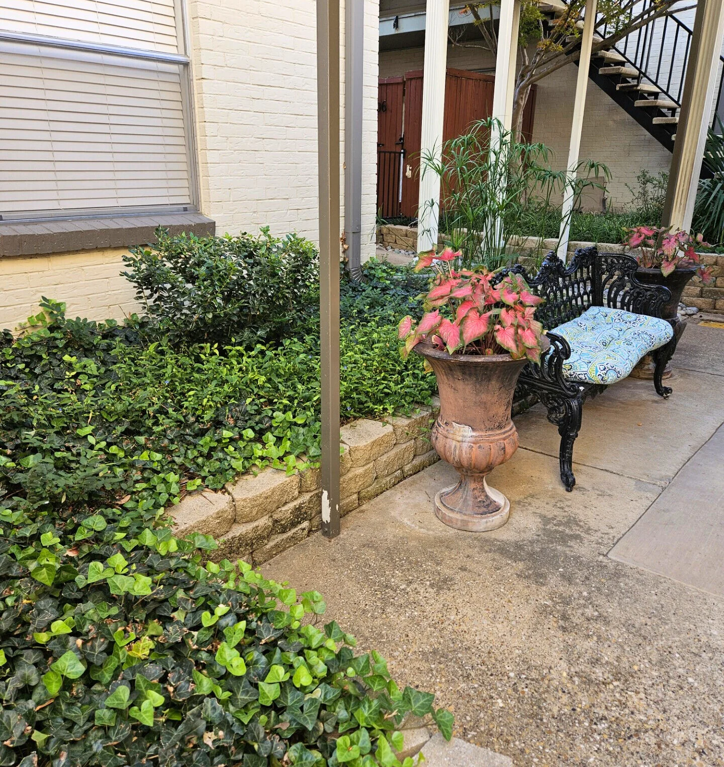 Small garden nook with a cushioned bench, a stone planter of pink flowers and green shrubs