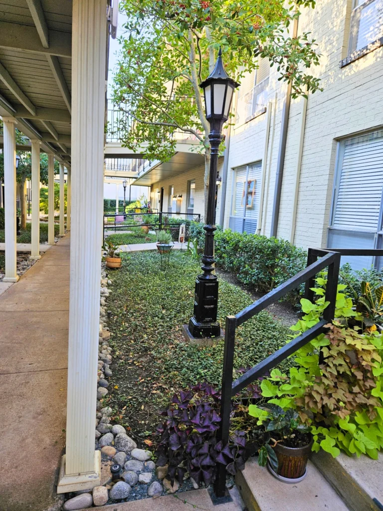 Covered walkway along a building with a black lamppost, an iron railing and flowering plants