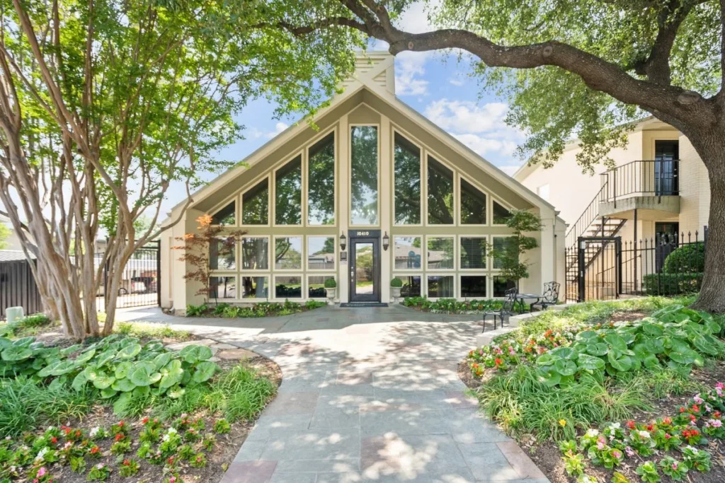 Glass-fronted building with a tall peaked roof, framed by trees and flower beds along a walkway