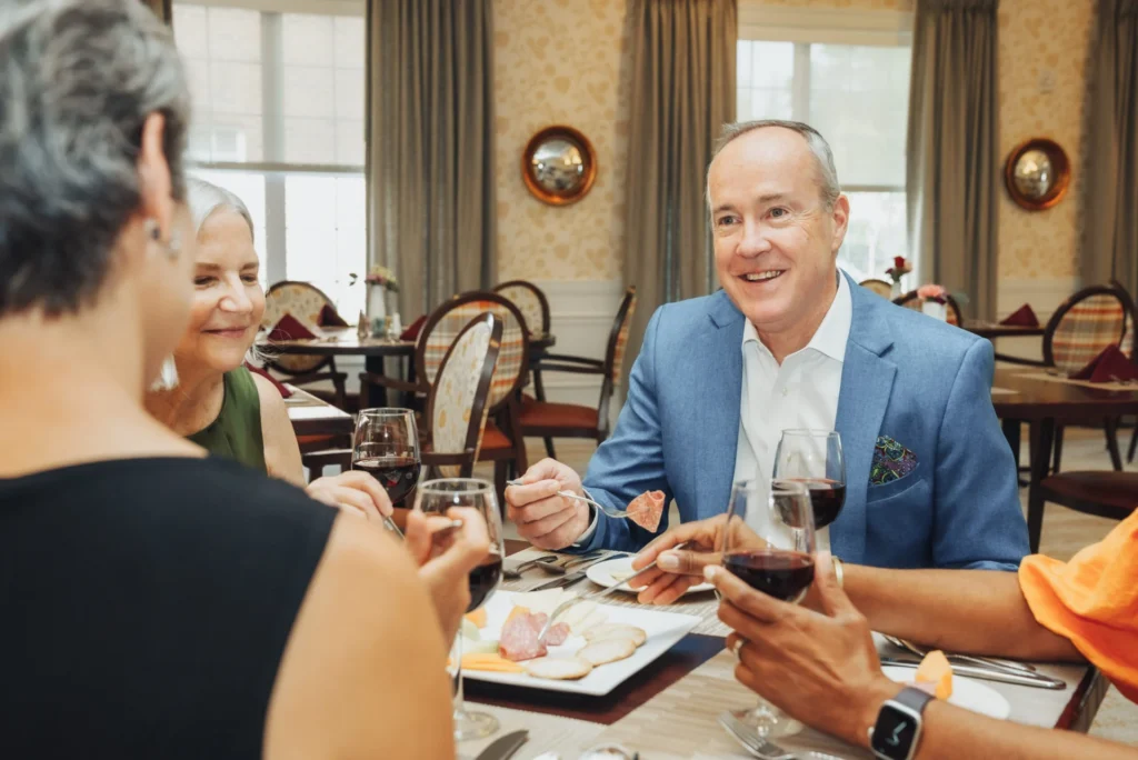 Smiling older man in a blue blazer holding a glass of red wine, seated at a dining table with friends