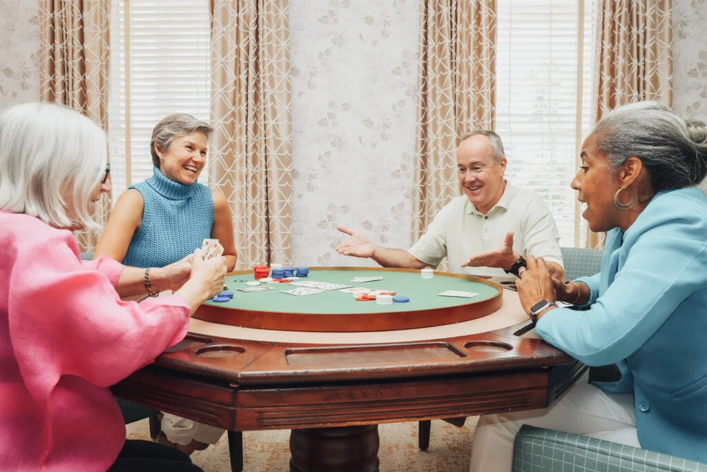 Older adults playing a card game around a round table in a living room with curtains