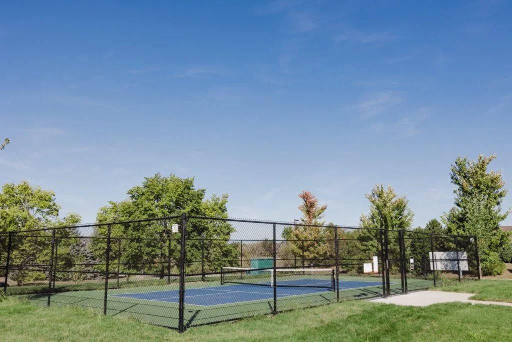 A fenced outdoor pickleball court in Hilliard, Ohio is surrounded by green grass and trees under a clear blue sky, offering luxury senior living amenities.