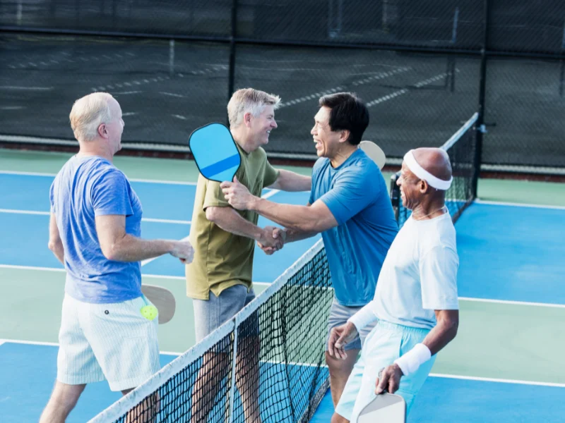 Older players shaking hands across the net on an outdoor court, holding paddles