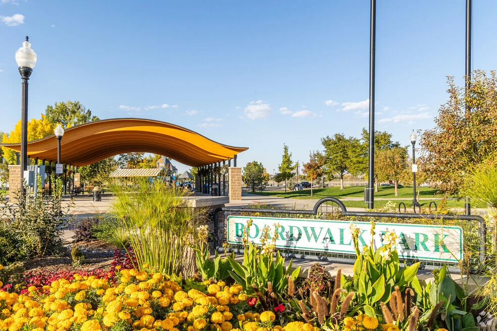 Flower beds and a park sign in front of a pavilion and lampposts under a blue sky