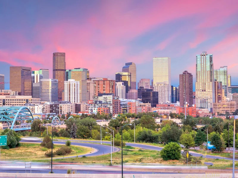 City skyline at sunset with pink clouds, seen across a park and a river