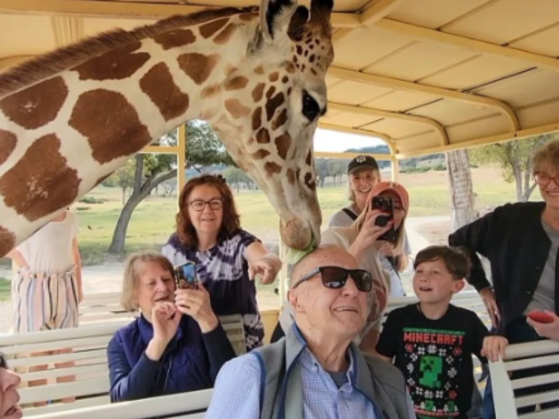 Group of visitors, including an older man, at a covered viewing deck as a giraffe leans in over the railing