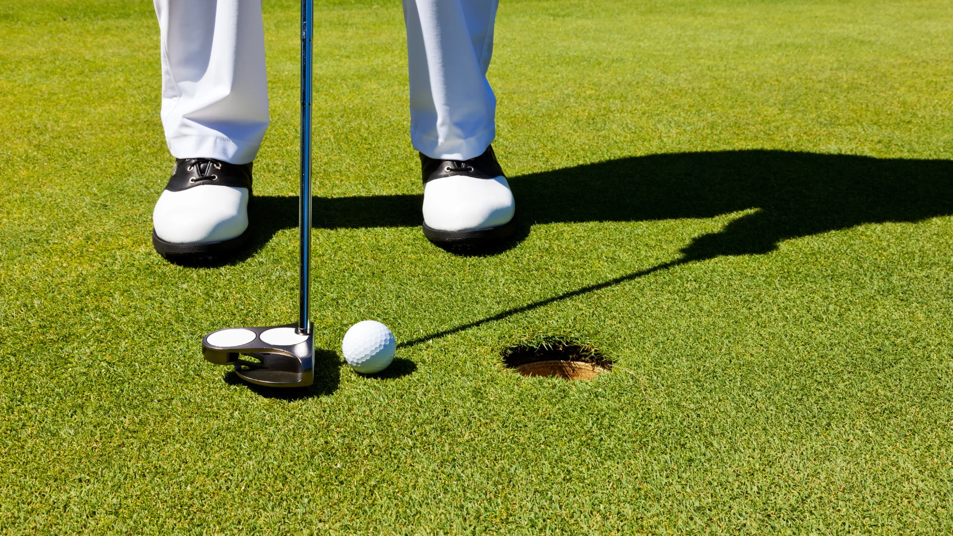 Close-up of a golfer's white shoes beside a golf ball at the edge of a hole on a putting green