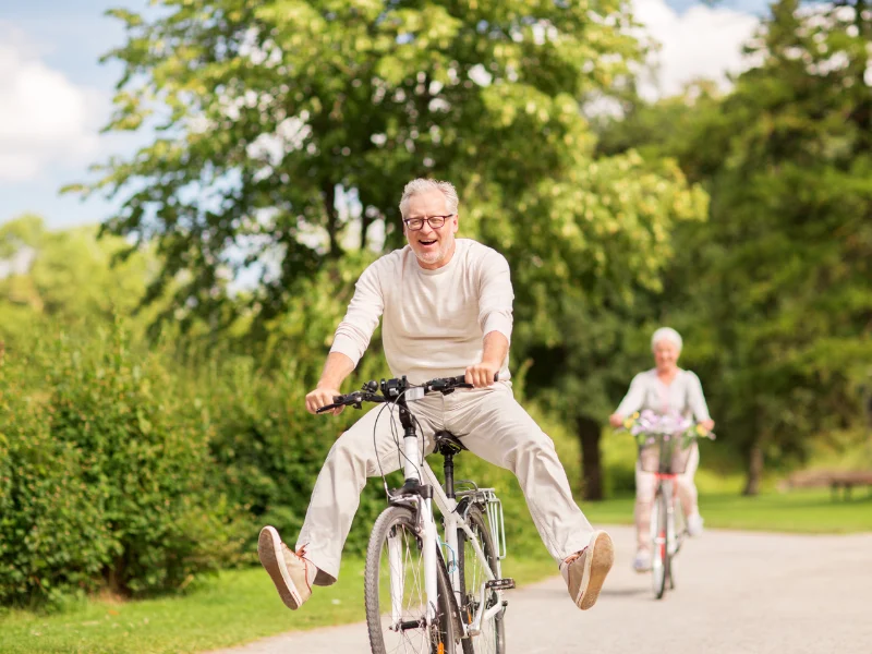 Older man smiling as he rides a bicycle on a tree-lined path, with another cyclist behind