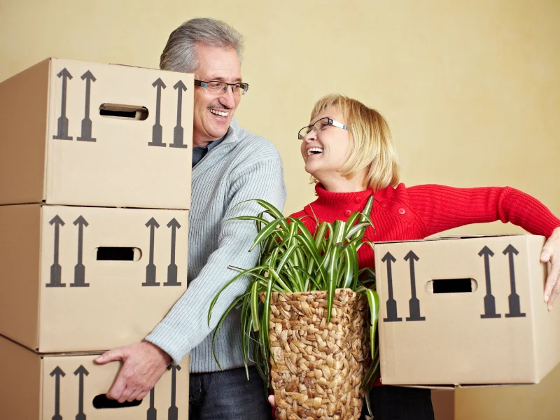 Older man and woman smiling as they carry moving boxes and a potted plant