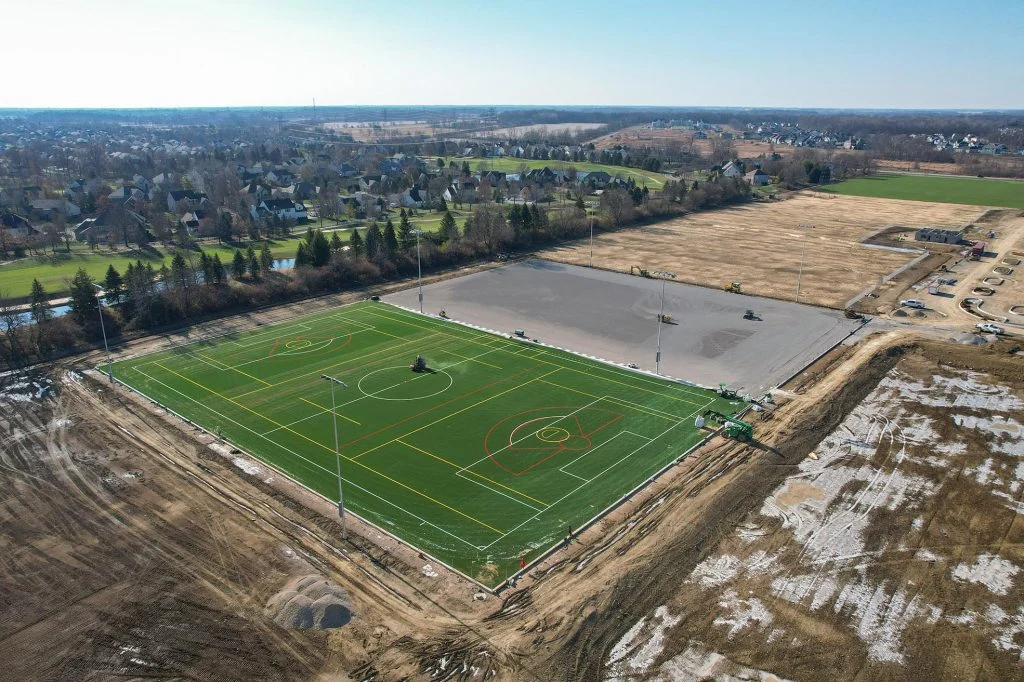 Aerial view of a green athletic field next to a graded construction site and surrounding neighborhoods