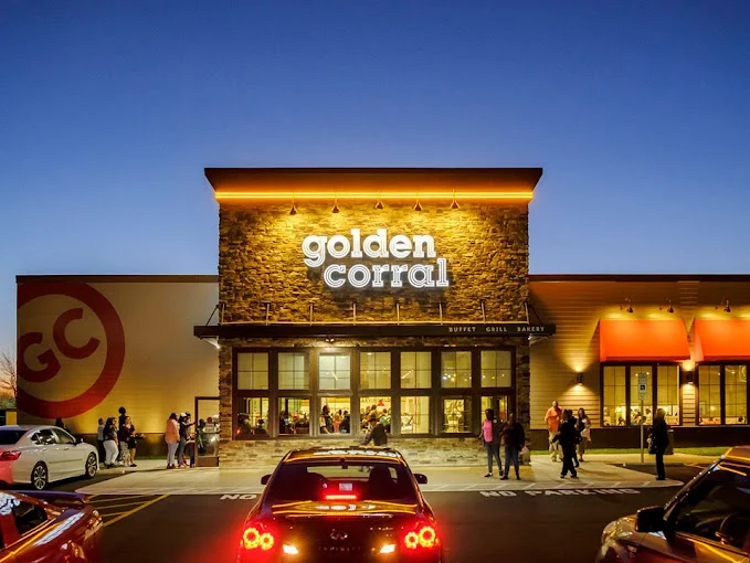 Restaurant entrance lit at dusk with a lit sign above the doors and cars in the lot