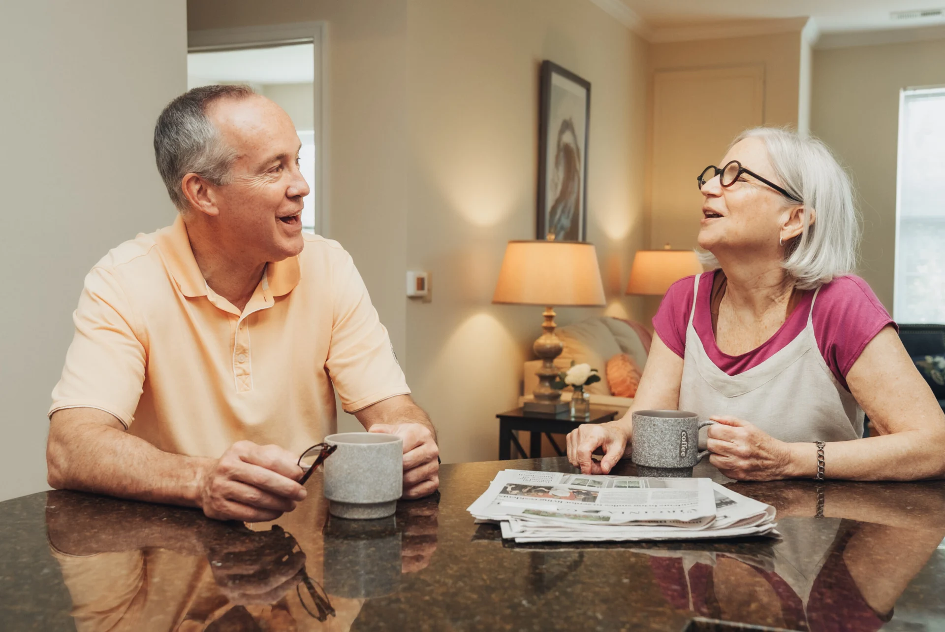 Older man and woman smiling over coffee and a newspaper at a table