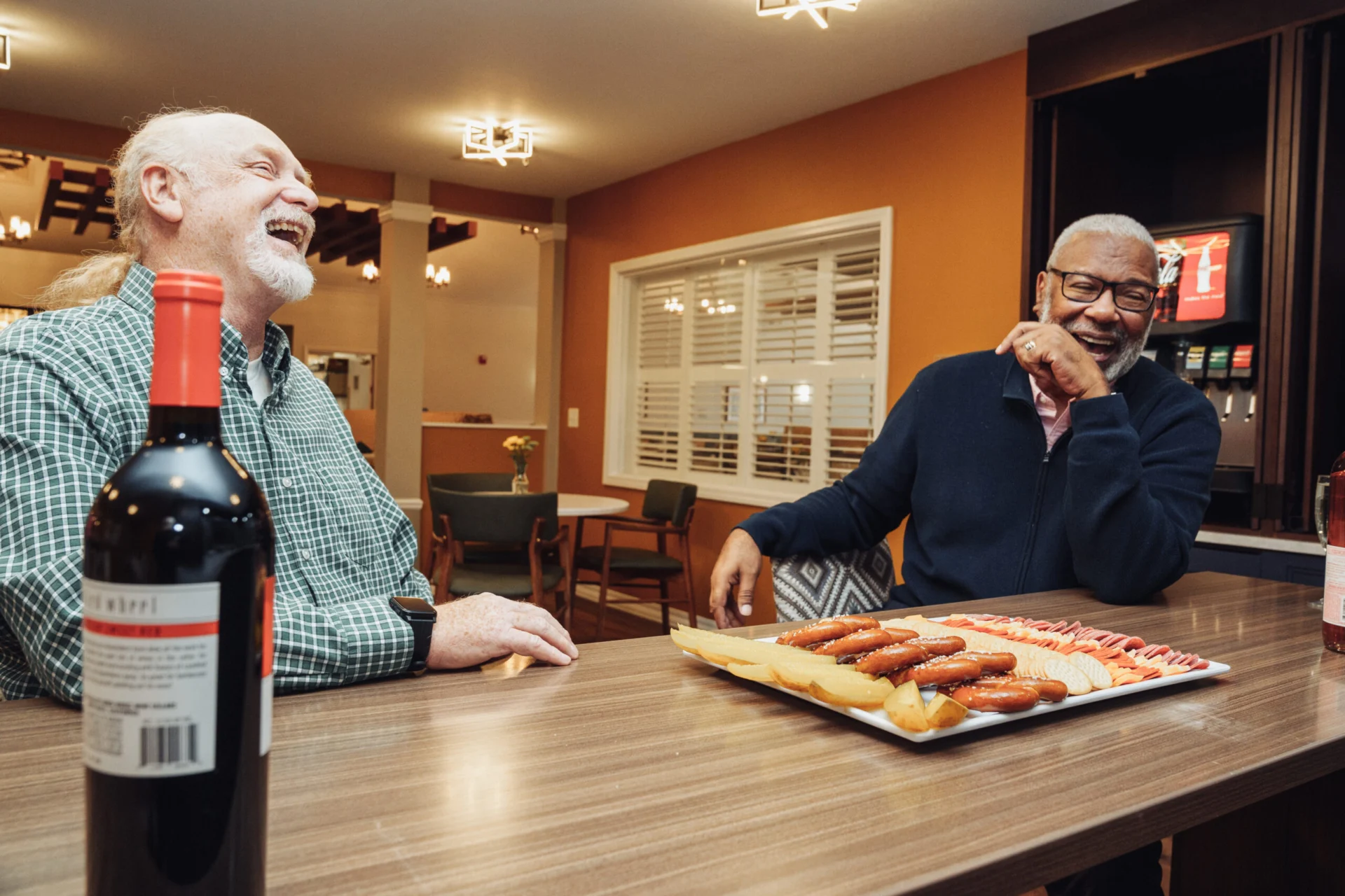 Two older men chatting at a wooden table with a bottle of red wine and a plate of appetizers