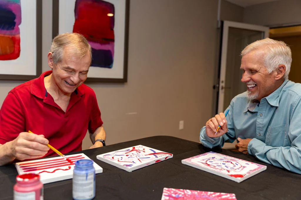 Two older men painting decorative pieces at a table, laughing