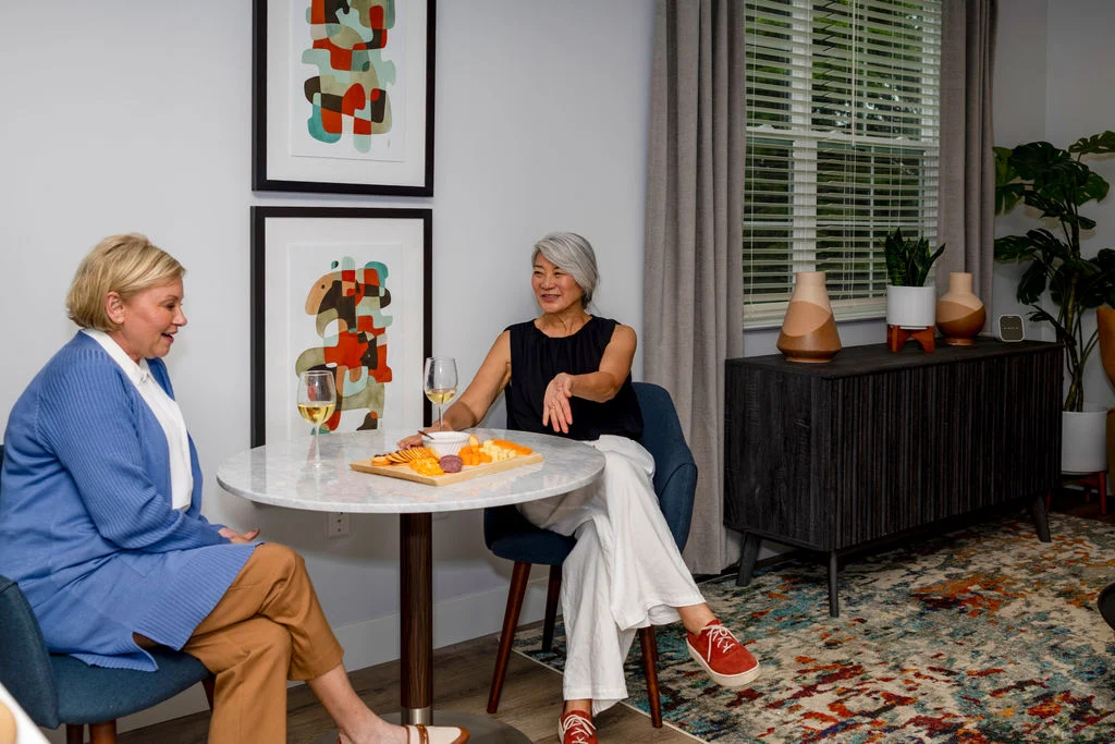 Two older women chatting at a small round table with a plate of pastries, with framed art on the wall
