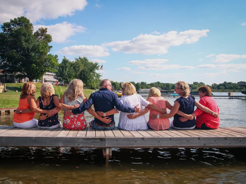 Group of friends seated on a wooden dock with their arms around each other, looking out at the water