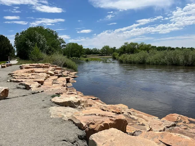 Rocky shoreline beside a calm river, with trees and a blue sky with clouds