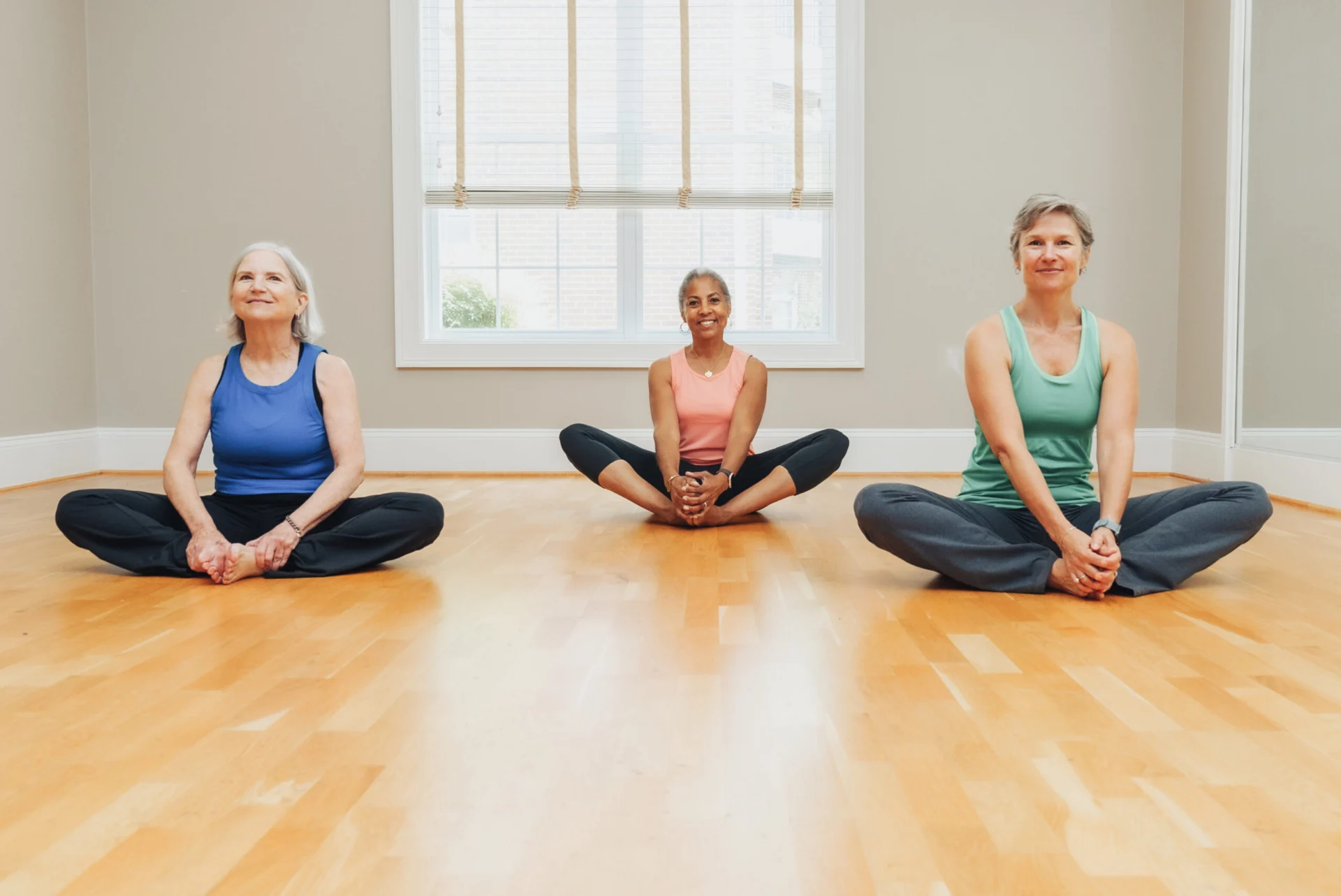 Three women sitting cross-legged on a wood floor in a bright studio, stretching