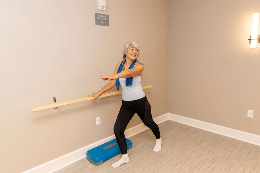 Woman with a towel around her neck stretching at a wall-mounted barre in a studio