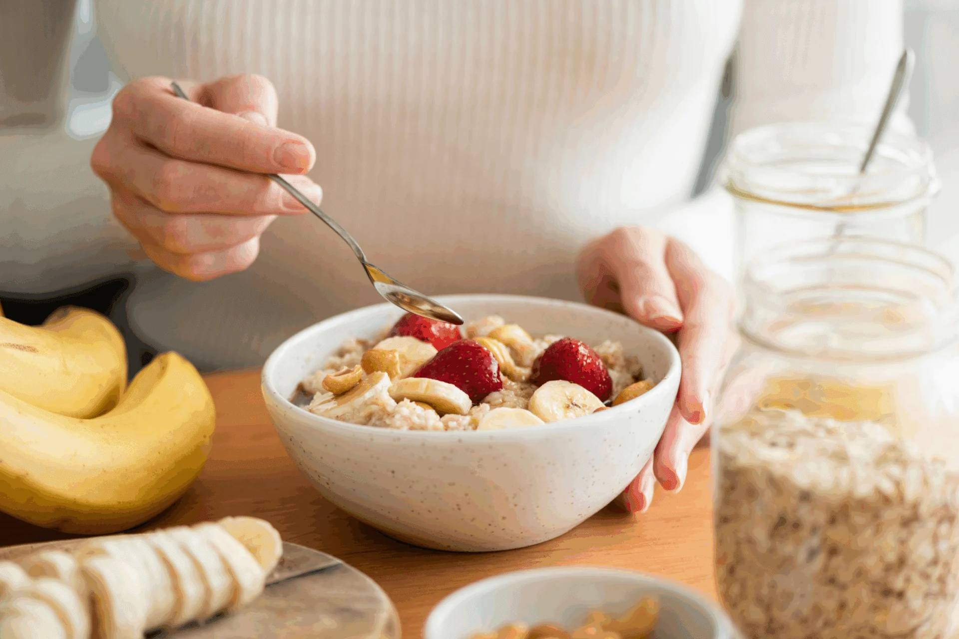 Hand holding a spoon over a bowl of granola and berries, with sliced bananas and jars of oats nearby