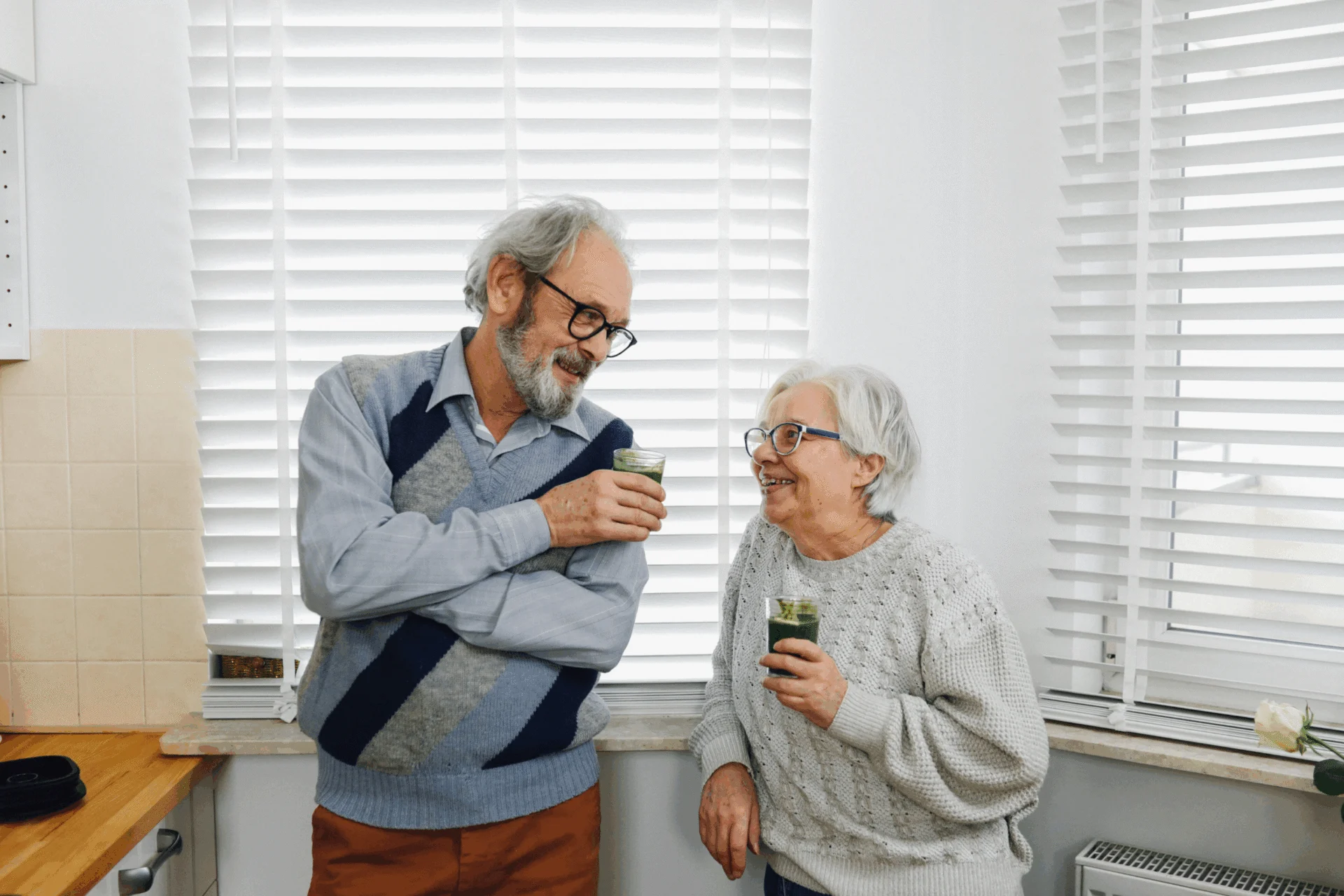 Older man and woman chatting in a kitchen while holding small glasses of green juice