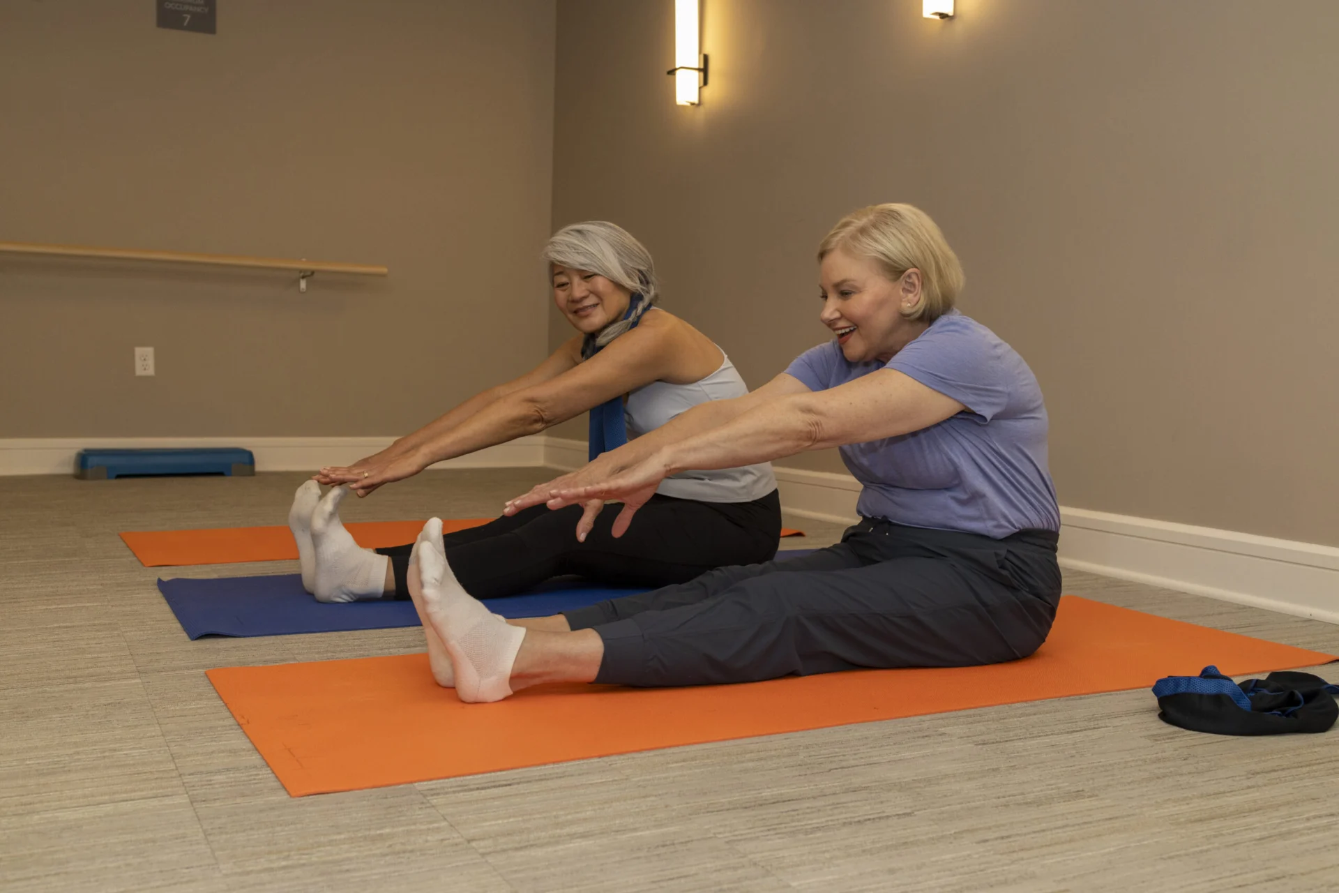 Two older women stretching while seated on mats in an exercise studio