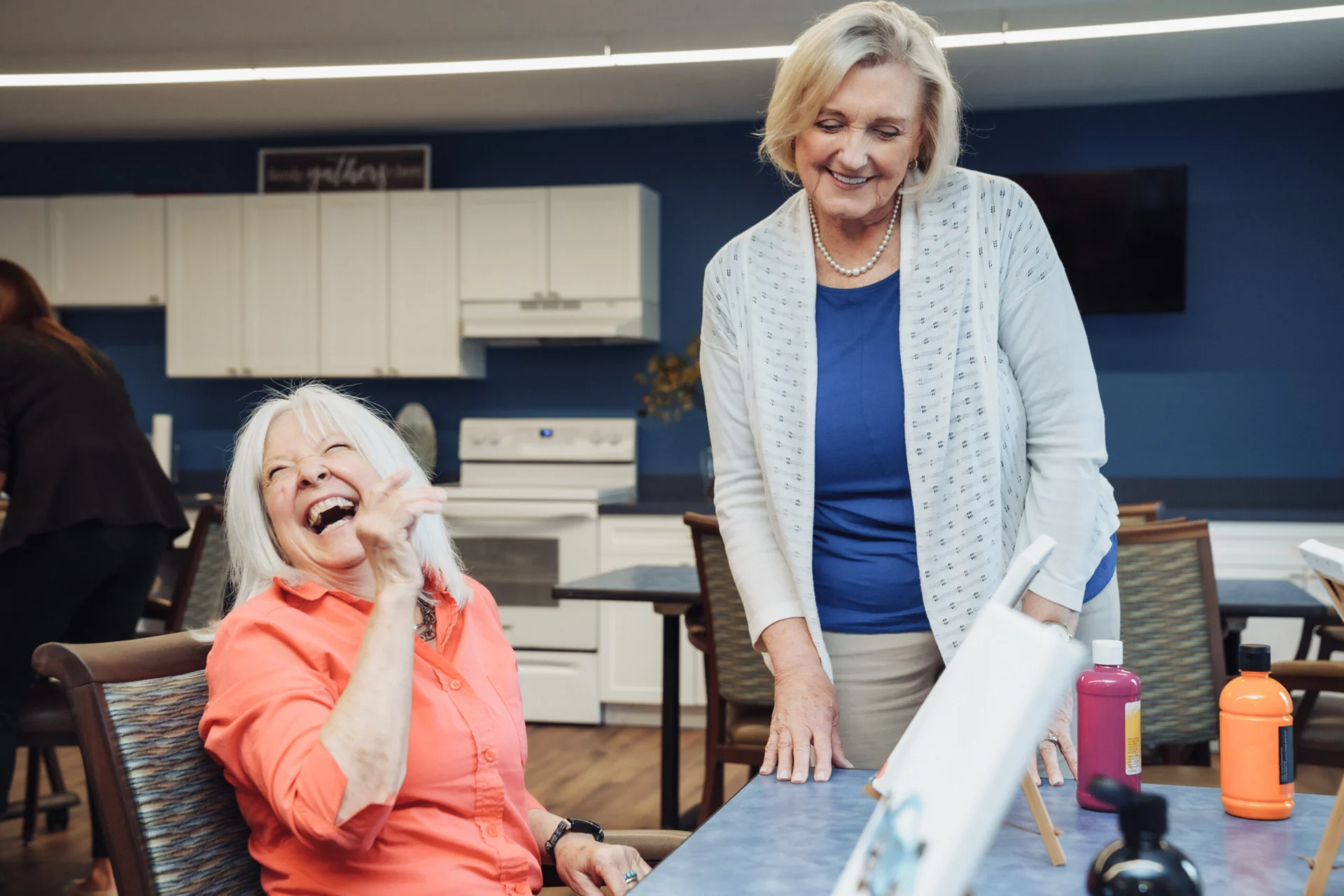 Older woman laughing at a table while another woman stands beside her smiling, in a bright kitchen