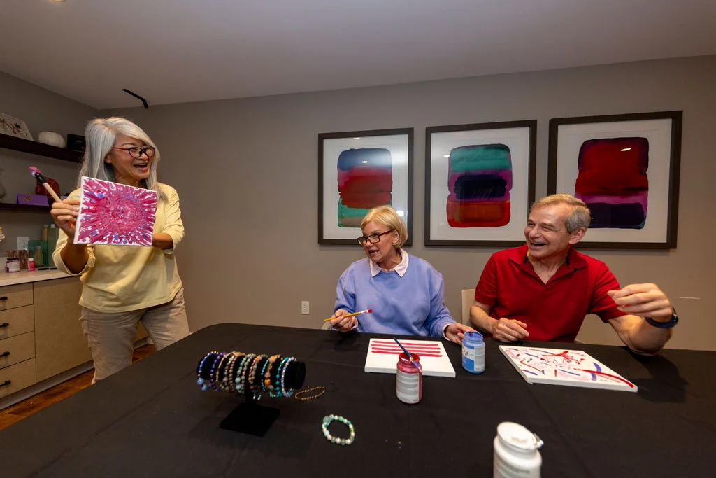 Older adults making bracelets at a dark table, with a woman standing and holding up a beaded piece