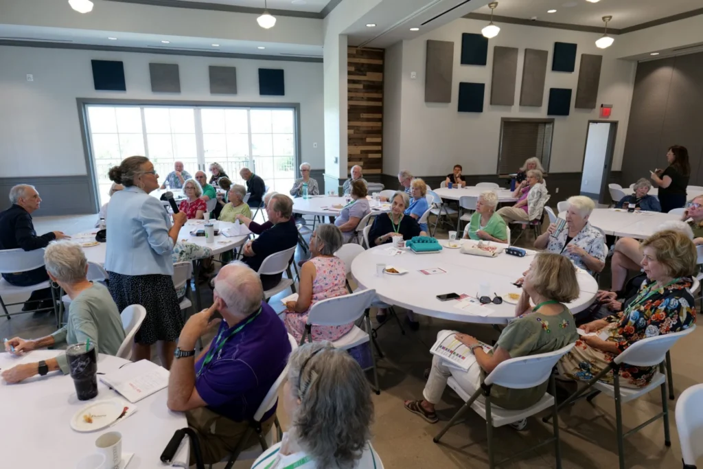 Large group of older adults seated at round tables in a community room during an event