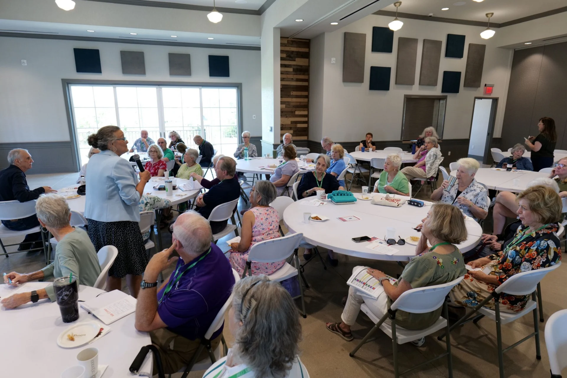 Large group of older adults seated at round tables in a community room during an event
