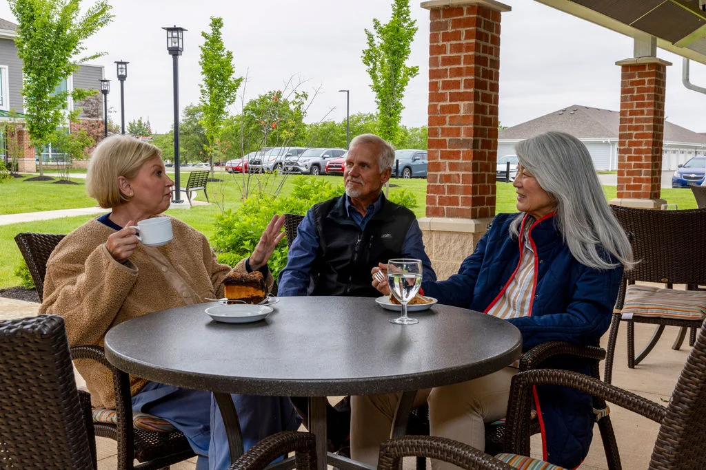 Three older adults chatting over coffee at a patio table beside brick columns