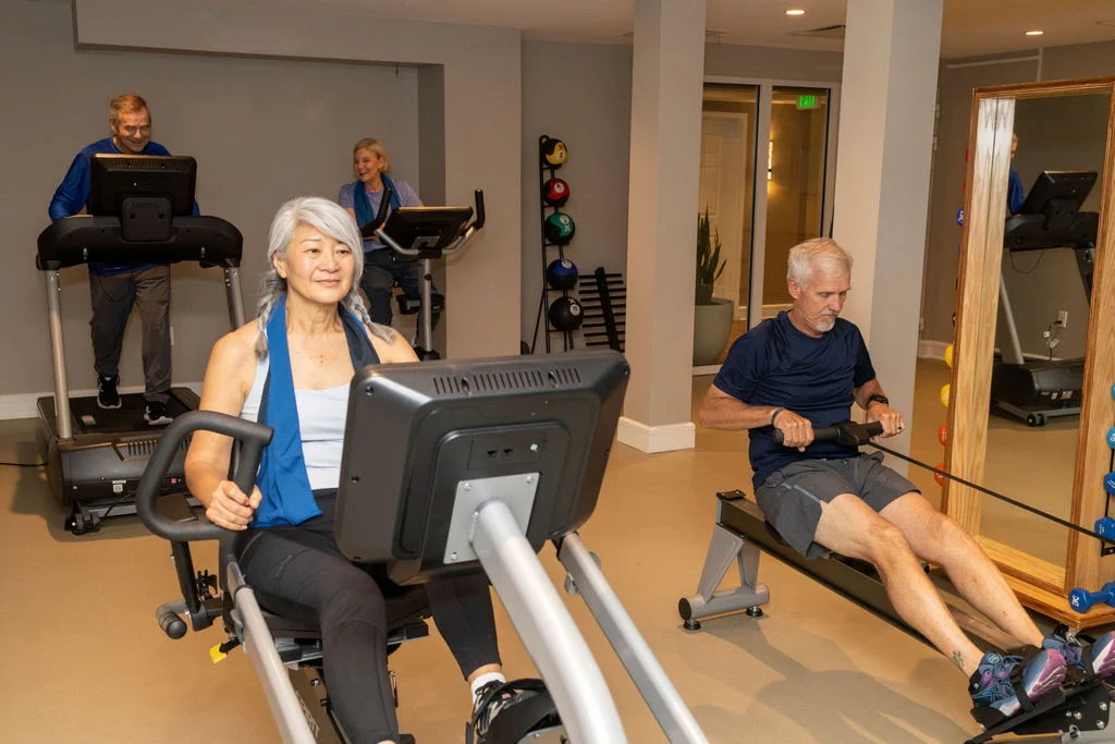 Older adults exercising on a stationary machine and a rowing machine in a fitness room, with other people on treadmills behind