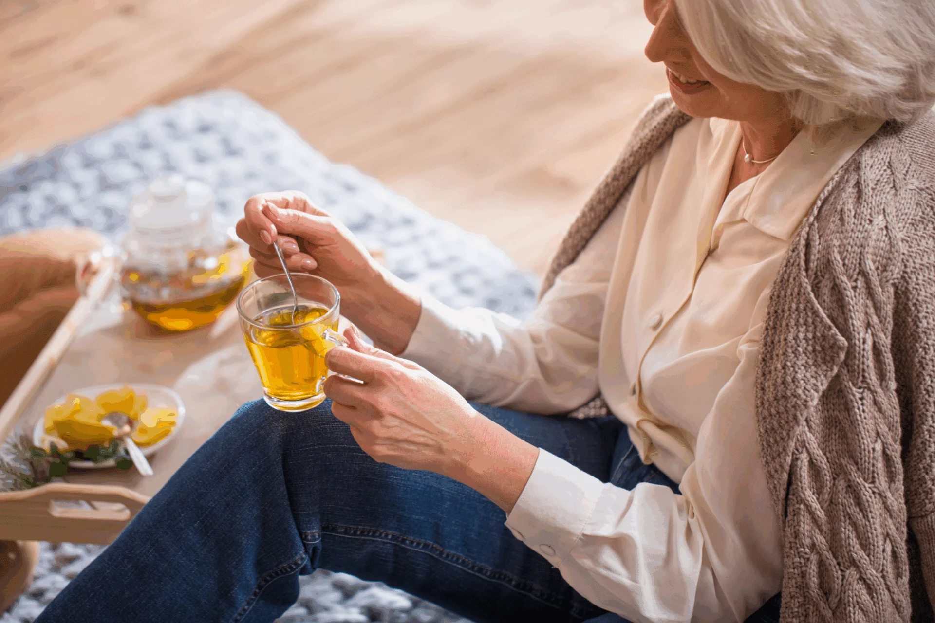 Older woman in a cardigan stirring a cup of tea at a table with a small tea set