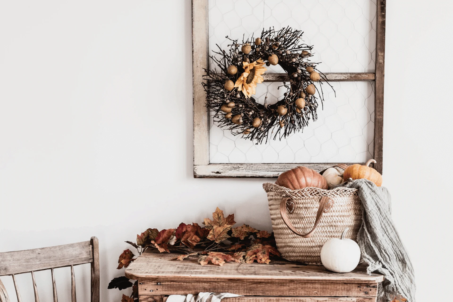Fall wreath in a rustic frame above a bench with a woven bag, dried leaves and a white pumpkin