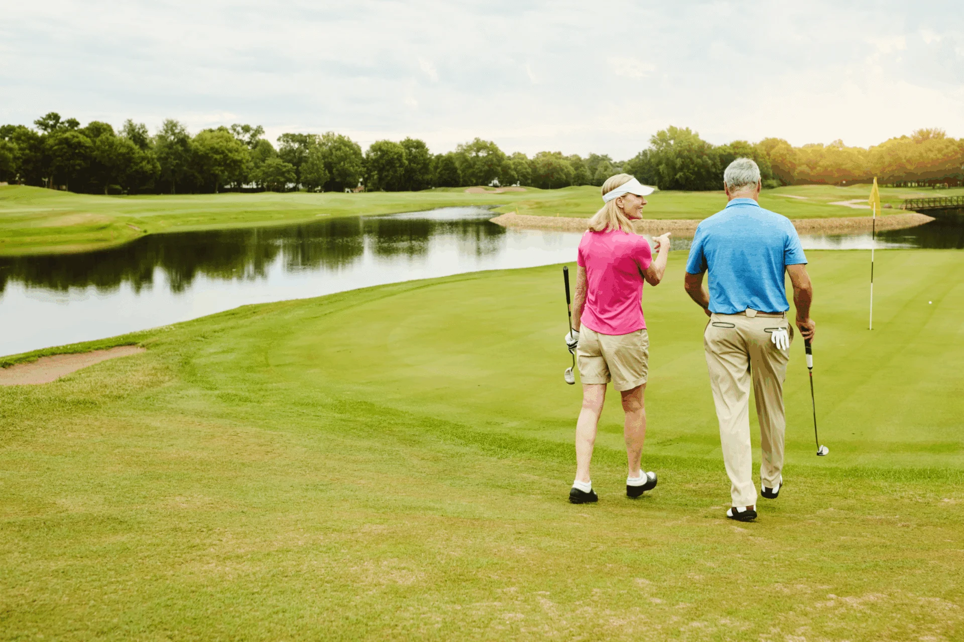 Two golfers walking on a fairway toward a pond with trees beyond