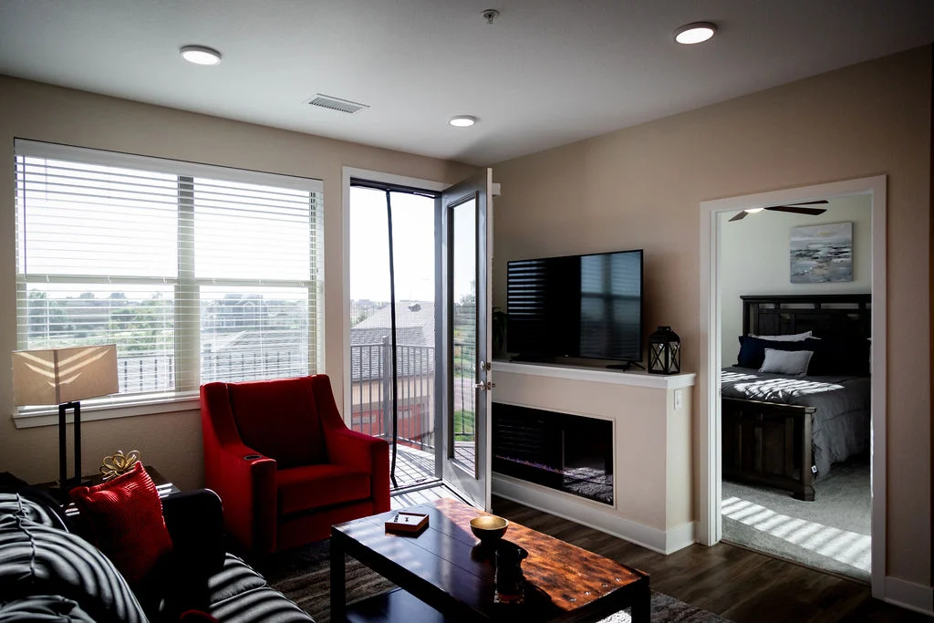 Apartment living room with a red armchair, a wall-mounted TV over a fireplace and a bedroom visible beyond