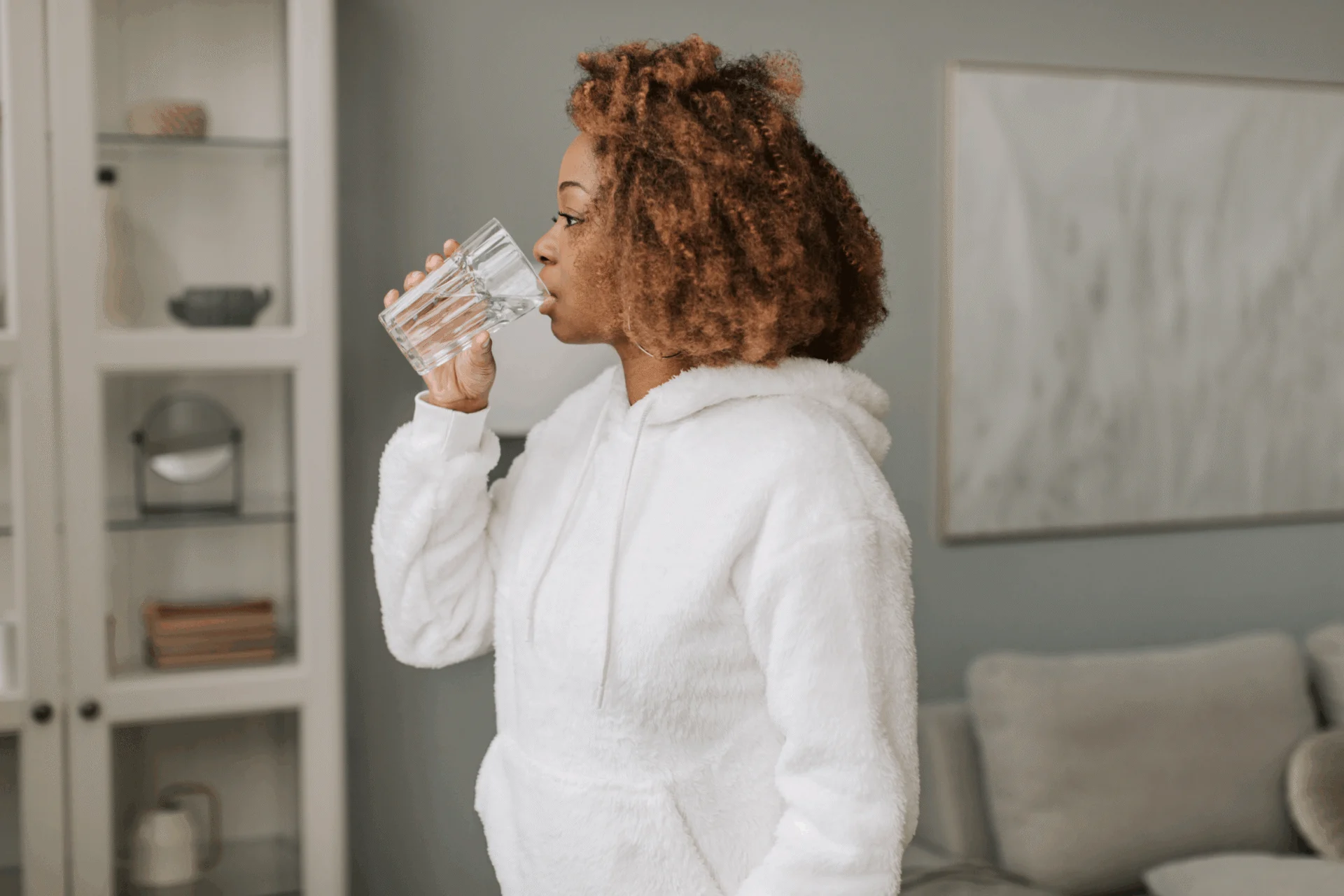 Woman in a white hoodie drinking a glass of water in a living room