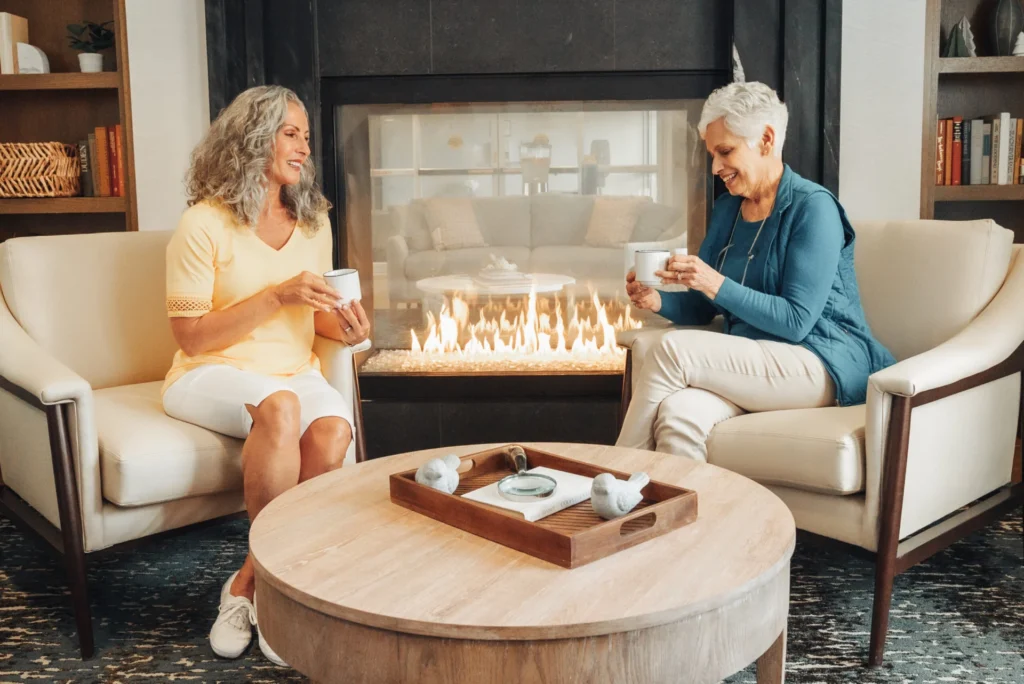 Two older women sipping tea in armchairs beside a gas fireplace, with a tray on a round table