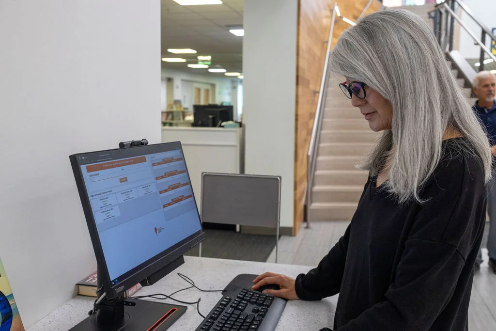 A woman with long gray hair and glasses sets small goals as she uses a desktop computer at a library counter. Stairs and bookshelves line the background, while another person walks by.