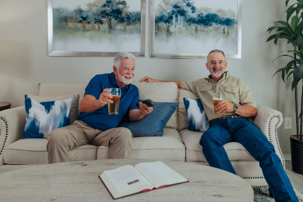 Two older men relaxing on a sofa holding drinks and smiling