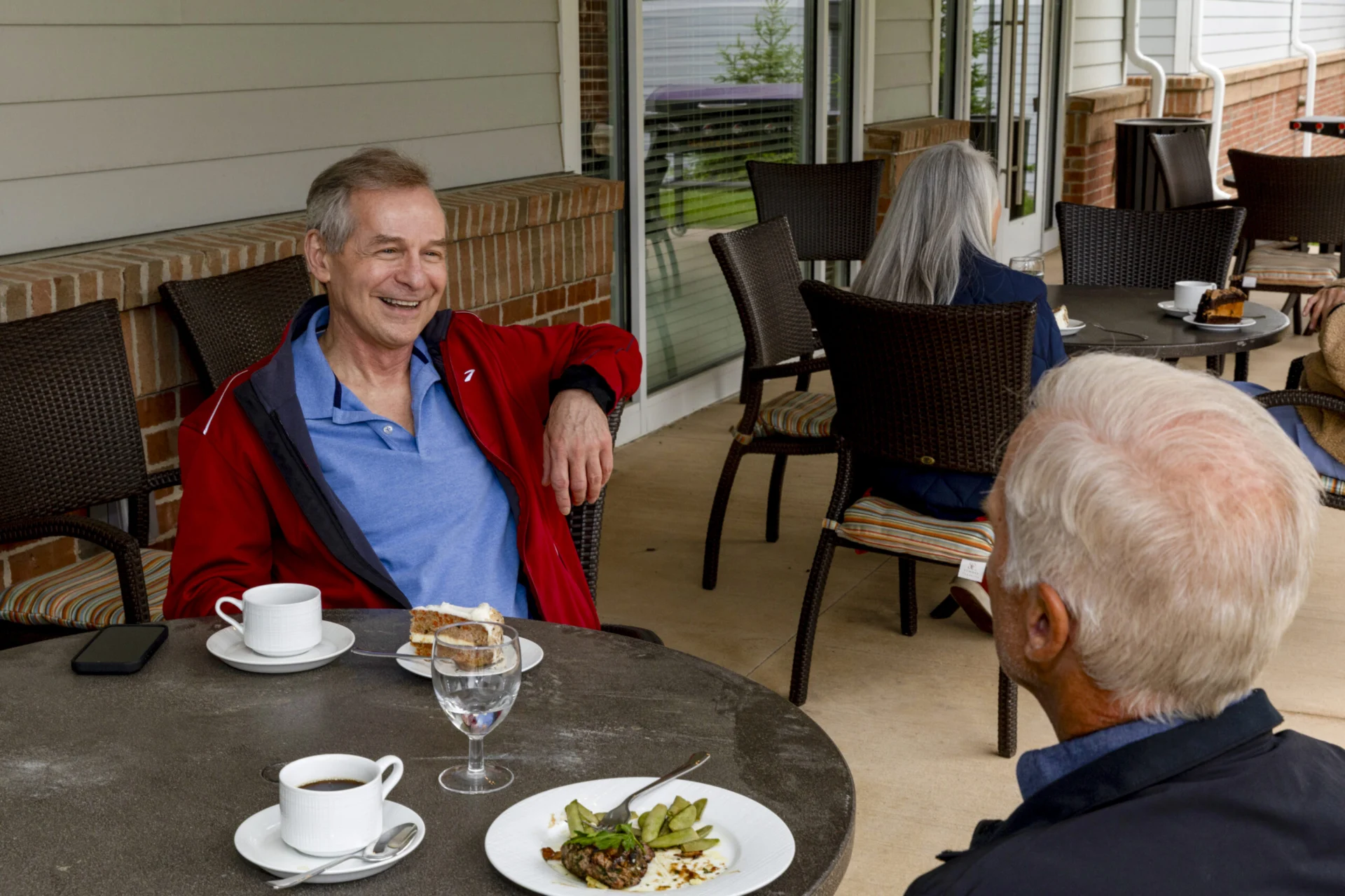 Two senior men sit at an outdoor café table in Hilliard, Ohio, smiling and conversing over coffee and dessert. Other patrons are visible nearby, enjoying the luxury of a relaxed afternoon beside a brick wall and large windows.
