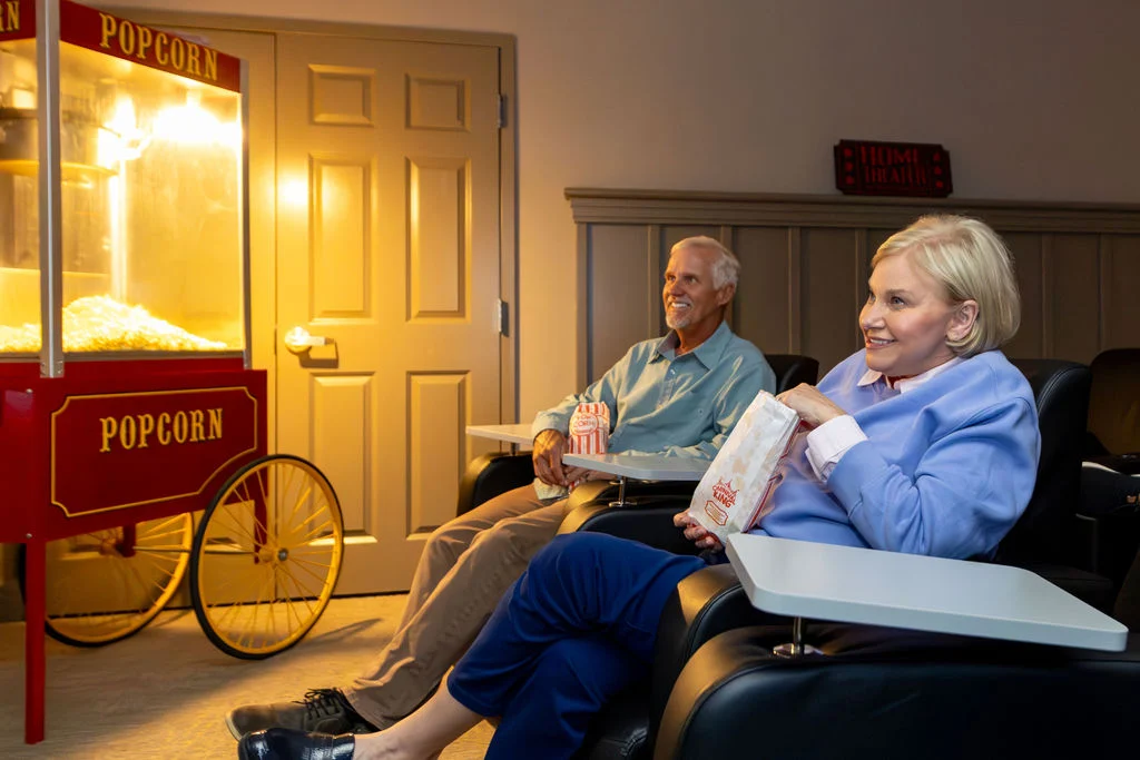Older man and woman relaxing in recliners with snacks, beside a red popcorn cart