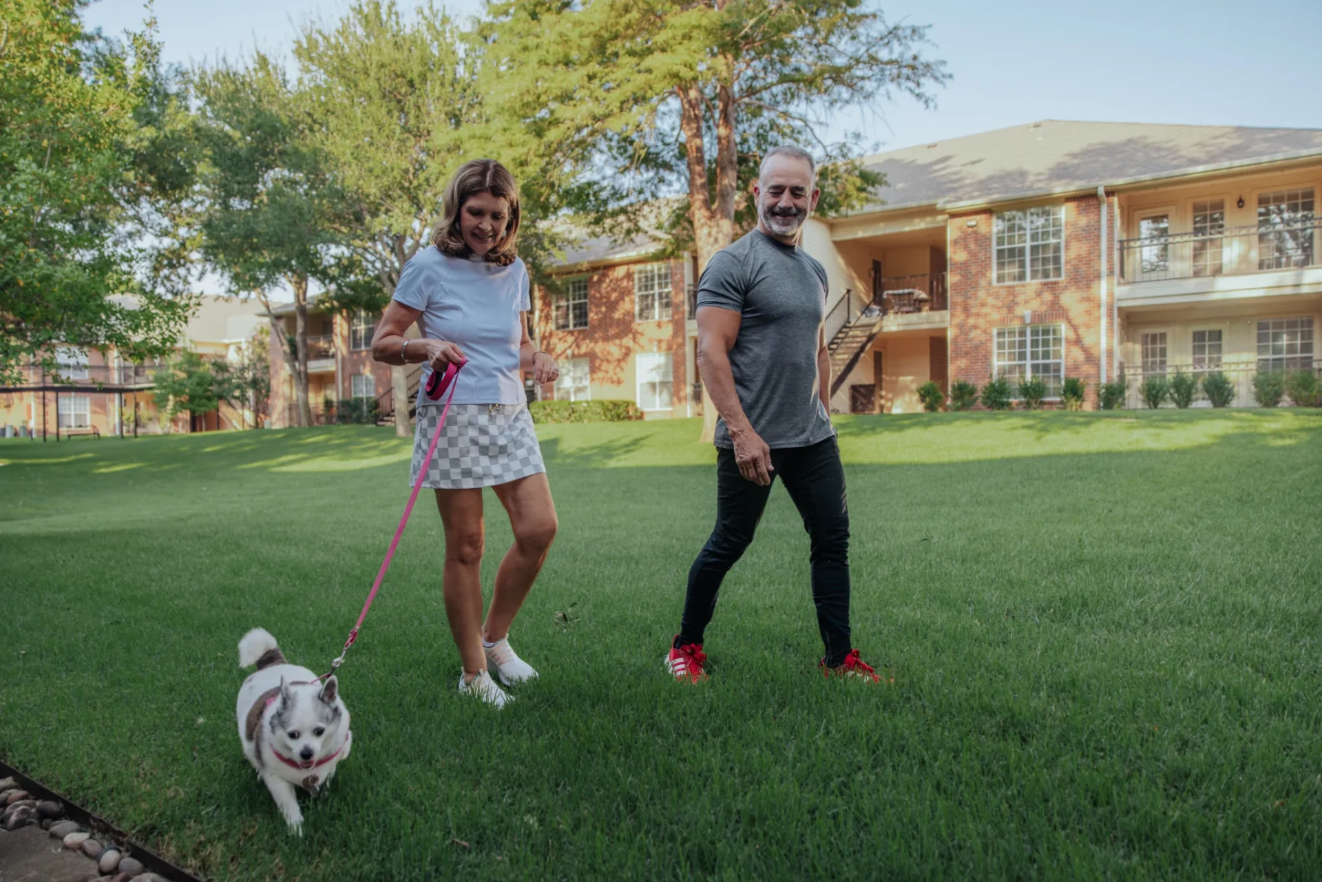 Couple walking a small dog on a leash across a lawn in front of apartment buildings