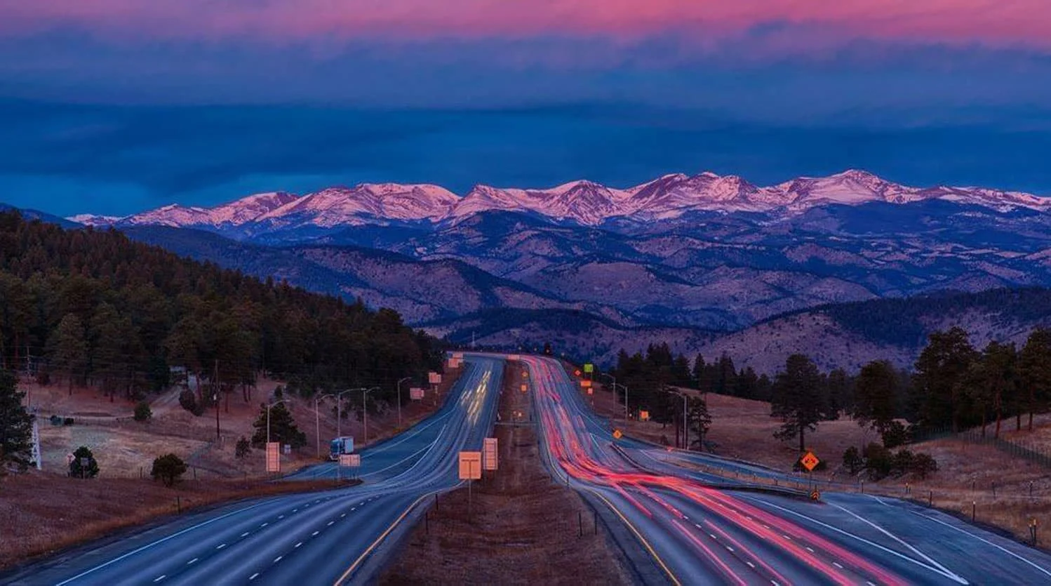 Highway with light trails leading toward snow-capped mountains at dusk