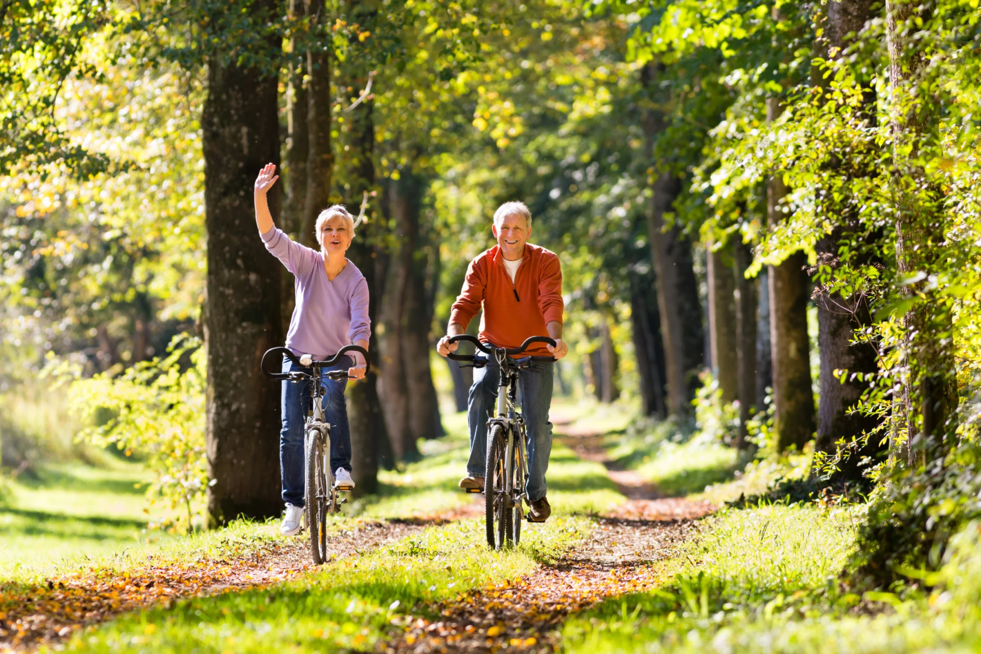 Two cyclists riding on a tree-lined trail with fallen leaves on the path