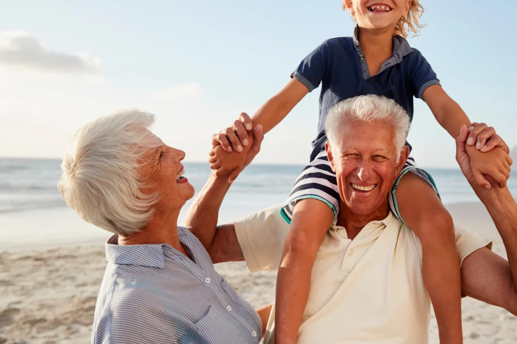 Older man carrying a child on his shoulders while a smiling older woman holds the child's hand, on a beach