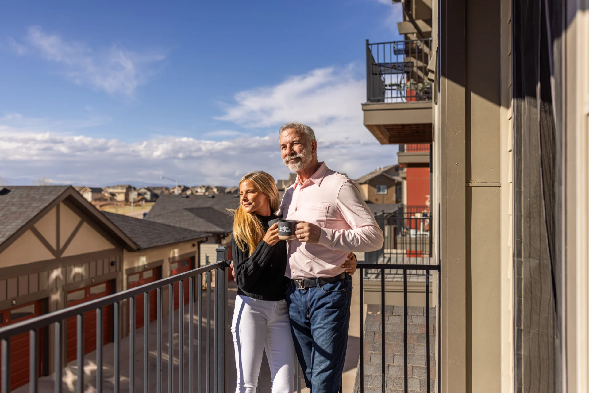 Couple standing on a balcony holding mugs, with rooftops and clouds behind them