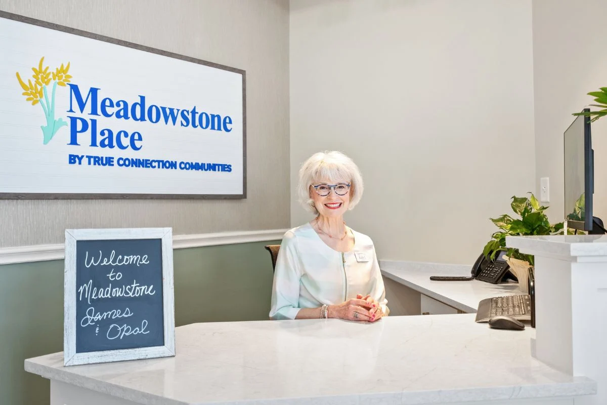 Smiling receptionist at a white front desk under a wall-mounted sign, with a small welcome chalkboard