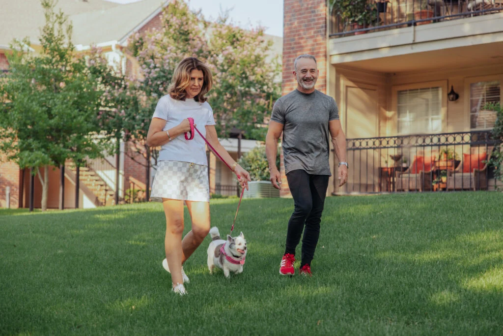 Couple walking two small dogs on leashes across a lawn beside brick apartment buildings