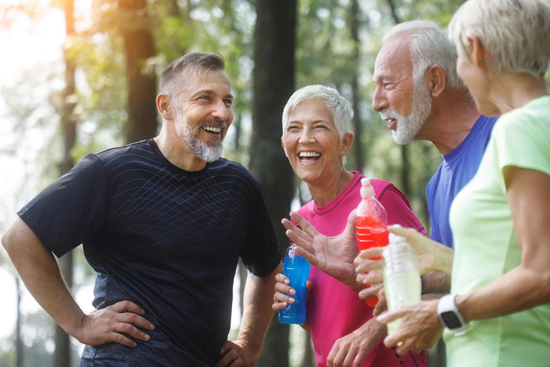 Four older adults in athletic clothing stand outdoors, smiling and laughing while holding water bottles at Verena at Hilliard, with trees and sunlight in the background. They appear to be enjoying a break from exercise.